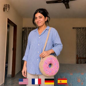 Könnte beinhalten: Eine runde, beige Häkeltasche mit rosa Zuckerguss und bunten Streuseln. Die Tasche hat einen beigen Riemen und wird über der Schulter getragen. Die Tasche ist wie ein Donut geformt.