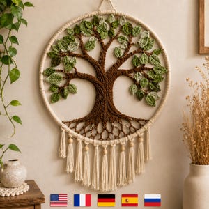 May include: A handcrafted wall hanging featuring a tree design. The tree has a brown trunk and branches with green leaves, set within a cream-colored circular frame. The design includes hanging tassels and wooden beads.