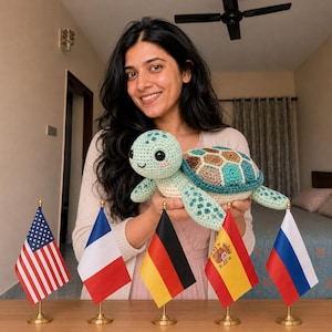 Op de afbeelding: Een lichtgroene gehaakte schildpad knuffel met een veelkleurig schild met bruine, blauwgroene en crèmekleurige zeshoekige patronen. De schildpad heeft grote zwarte veiligheidsogen, een geborduurde glimlach en roze blosjes op de wangen, vastgehouden voor vijf kleine vlaggetjes.