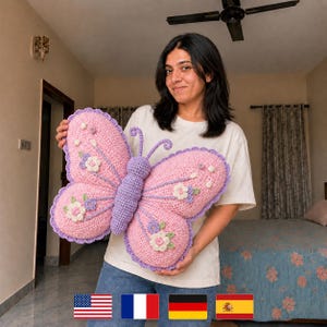 May include: A large, handmade crochet butterfly pillow in shades of pink and purple. The butterfly has detailed wings with floral accents and a textured body. The pillow is designed with a scalloped edge and measures approximately 30 inches across.