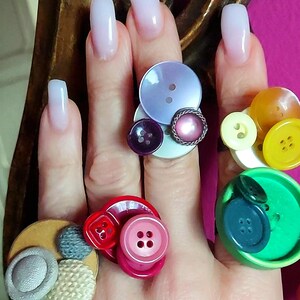 Può includere: Una collezione di anelli con gruppi di bottoni sovrapposti in vari colori, tra cui viola, giallo, verde, rosso, rosa e beige. I bottoni variano per dimensioni, consistenza e finitura, alcuni con rivestimento in tessuto e altri realizzati in plastica liscia.