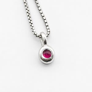 May include: A silver pendant necklace featuring a small, round, bezel-set vibrant pink gemstone. The pendant hangs from a delicate silver box chain. The metal has a polished finish, and the gemstone is centered within the circular silver frame.