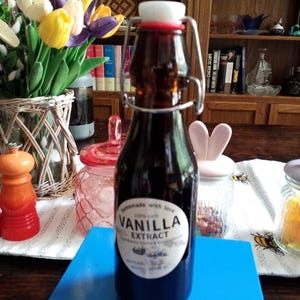 May include: A dark amber glass bottle with a swing-top closure and a white ceramic stopper. The bottle features a round white label with the text "Homemade with love, 100% pure, VANILLA EXTRACT, Ingredients: Vodka & Vanilla Beans, BOTTLED: 08/2023".