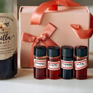 May include: A collection of vanilla extract products. A large glass bottle of homemade vanilla extract is next to four small bottles labeled "Hawaii", "Tahiti", "Uganda", and "Mix". A pink gift box with a ribbon is also present.