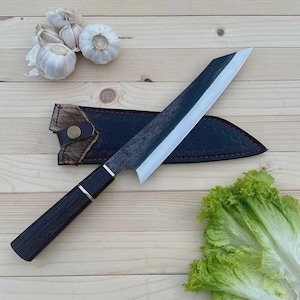 May include: A Japanese-style chef's knife with a dark wooden handle and a silver blade, resting on a wooden surface. The knife is accompanied by a black leather sheath, garlic, and lettuce.