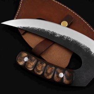 May include: A curved blade knife with a dark, textured metal finish along the spine and a polished silver edge. The handle is crafted from dark, grained wood with four distinct finger grooves and two silver rivets, resting on a brown leather sheath.