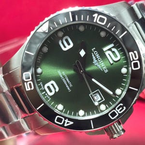 Pode incluir: Um relógio de pulso Longines em aço inoxidável com mostrador verde, bisel rotativo preto e ponteiros prateados. O mostrador apresenta o logótipo Longines, grandes numerais árabes às 6, 9 e 12, uma janela de data às 3 e Automatic 30bar 300meter.