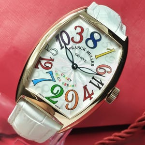 Puede incluir: Un reloj de pulsera Franck Muller Geneve con caja dorada en forma de tonel y correa de piel blanca con textura de cocodrilo. La esfera blanca presenta grandes números arábigos multicolores, manecillas negras y el texto "Crazy Color Dreams" y "Swiss Made".
