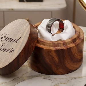 May include: A wooden ring box with the words "Eternal Promise" engraved on the lid. Inside, a ring with a dark, patterned exterior and a red interior rests on white fabric. The box is open, revealing the ring.
