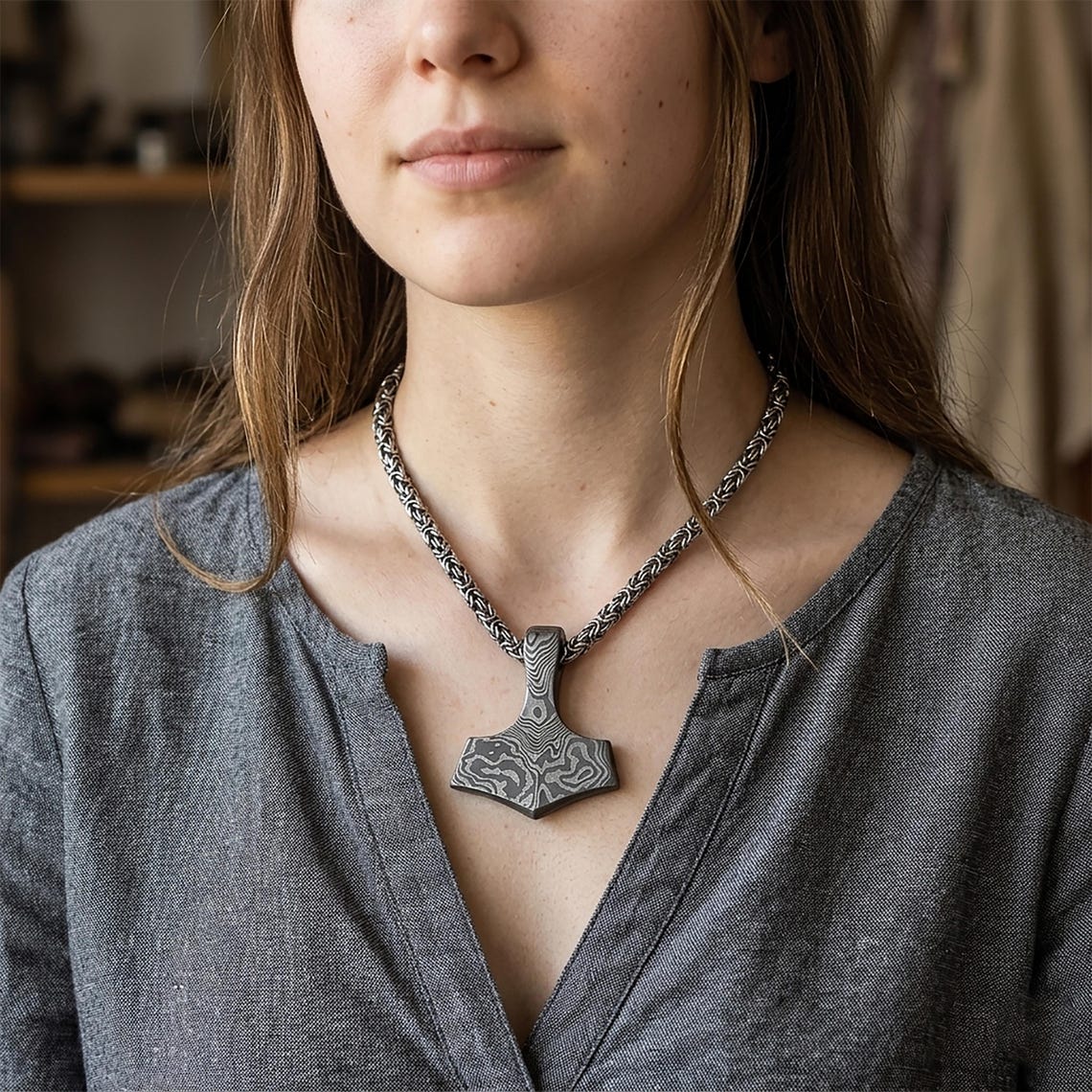 May include: A silver-colored pendant necklace featuring a hammer-shaped pendant with a patterned design. The necklace has a chain-link style and is worn on a person wearing a gray shirt.