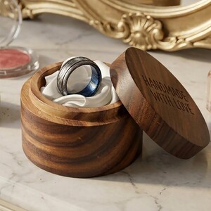 May include: A Damascus steel ring with a blue interior rests in a wooden ring box lined with white fabric. The box is open, with the lid nearby, and the text "HANDMADE WITH LOVE" is engraved on the lid.