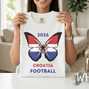 May include: A white t-shirt featuring a graphic of a butterfly with wings patterned like the Croatian flag. Above the butterfly is the text 2026, and below the butterfly is the text CROATIA FOOTBALL in red and blue lettering on the front center.