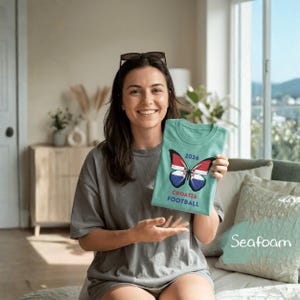 May include: A seafoam green t-shirt featuring a graphic of a butterfly with wings patterned like the Croatian flag. Above the butterfly is the text 2026, and below the butterfly is the text CROATIA FOOTBALL in a bold, sans-serif font.