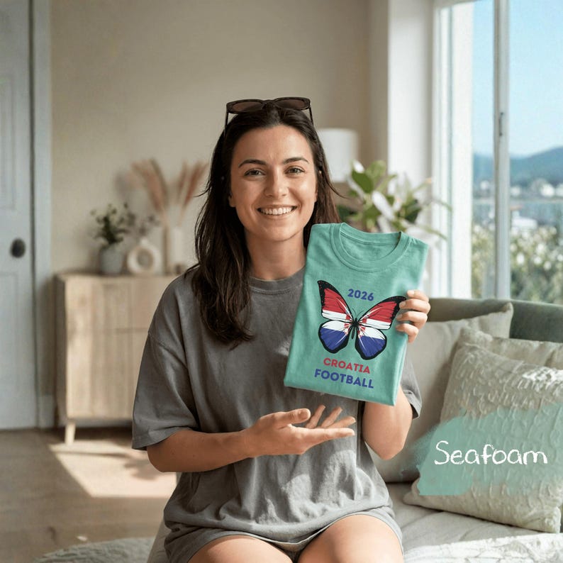 May include: A seafoam green t-shirt featuring a graphic of a butterfly with wings patterned like the Croatian flag. Above the butterfly is the text 2026, and below the butterfly is the text CROATIA FOOTBALL in a bold, sans-serif font.