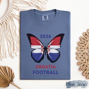 May include: A blue jean colored t-shirt featuring a butterfly graphic with the Croatian flag pattern on its wings. Above the butterfly is the text 2026, and below it is the text CROATIA FOOTBALL in red and blue lettering. The shirt has a crew neck.