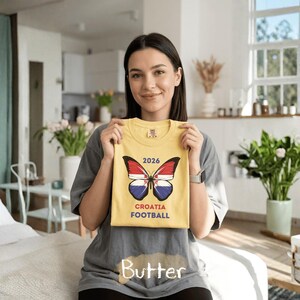 May include: A yellow t-shirt featuring a butterfly graphic with wings patterned like the Croatian flag. Above the butterfly is the text 2026, and below it is the text CROATIA FOOTBALL. The shirt is held up against a gray t-shirt background.