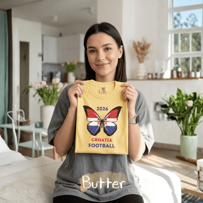 May include: A yellow t-shirt featuring a butterfly graphic with wings patterned like the Croatian flag. Above the butterfly is the text 2026, and below it is the text CROATIA FOOTBALL. The shirt is held up against a gray t-shirt background.