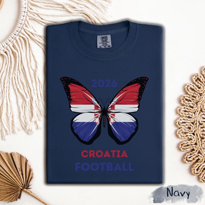 May include: A navy blue t-shirt featuring a butterfly graphic with wings patterned after the Croatian flag. The text 2026 is printed above the butterfly, while the words CROATIA FOOTBALL are printed below in red and blue lettering.