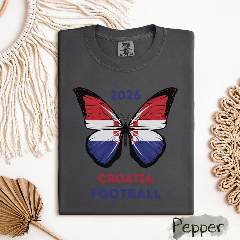 May include: A charcoal gray t-shirt featuring a butterfly graphic with wings patterned after the Croatian flag. Above the butterfly is the text 2026 in blue, and below it is the text CROATIA in red and FOOTBALL in blue. The shirt has a crew neckline.