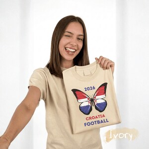 May include: An ivory t-shirt featuring a graphic of a butterfly with wings patterned like the Croatian flag. Above the butterfly is the text 2026, and below it is the text CROATIA FOOTBALL in red and blue lettering on the front of the garment.