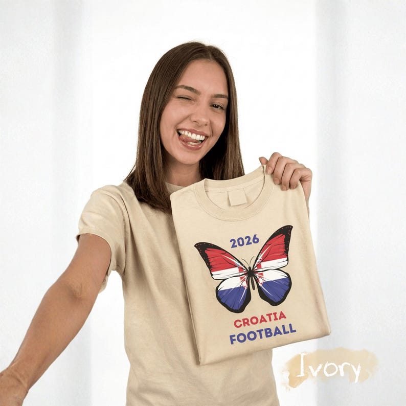 May include: An ivory t-shirt featuring a graphic of a butterfly with wings patterned like the Croatian flag. Above the butterfly is the text 2026, and below it is the text CROATIA FOOTBALL in red and blue lettering on the front of the garment.