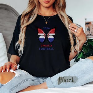 May include: A black t-shirt featuring a butterfly graphic with wings patterned like the Croatian flag. Above the butterfly is the text 2026, and below it is the text CROATIA FOOTBALL in blue and red lettering. The shirt is worn with light blue jeans.