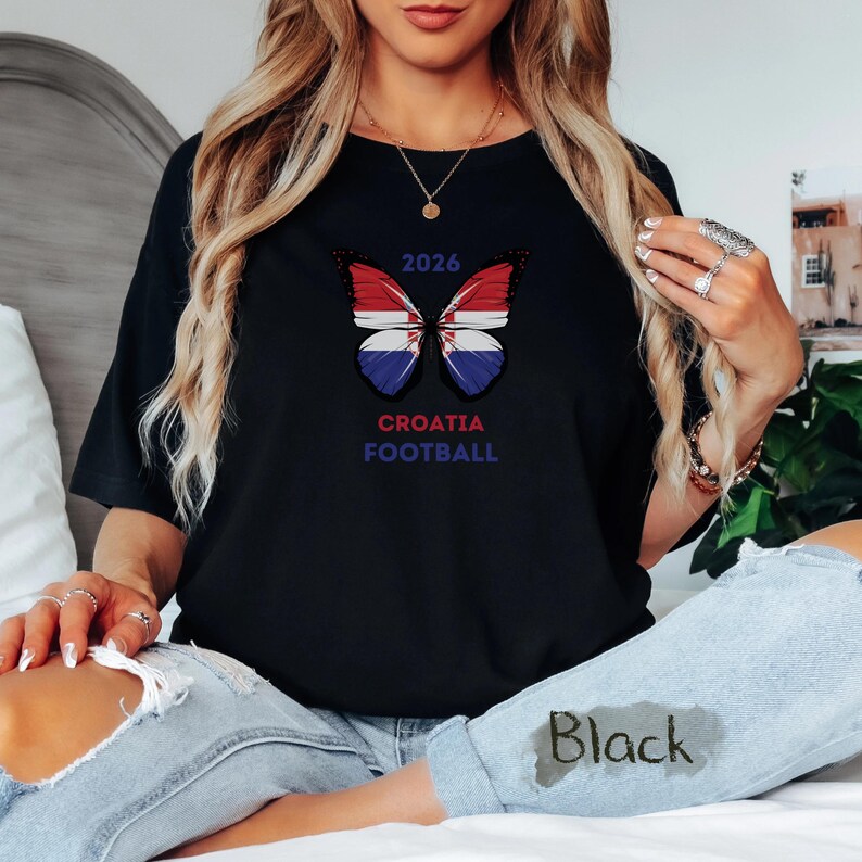 May include: A black t-shirt featuring a butterfly graphic with wings patterned like the Croatian flag. Above the butterfly is the text 2026, and below it is the text CROATIA FOOTBALL in blue and red lettering. The shirt is worn with light blue jeans.