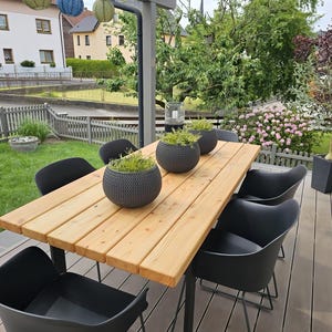 Könnte beinhalten: Ein rechteckiger Esstisch für den Außenbereich mit einer hellbraunen Holzplankenplatte und einem schwarzen Metallrahmen, dazu sechs schwarze Stühle aus geformtem Kunststoff. Drei dunkelgraue, kugelförmige, strukturierte Pflanzgefäße mit Grün.