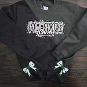 Könnte beinhalten: Ein dunkelgrünes Langarm-Sweatshirt mit dem Schriftzug POWERHOUSE Cheer aus weißen Strasssteinen auf der Brust. Das untere Ende jedes Ärmels ist mit einer weißen und grünen Schleifengrafik verziert. Ein Etikett mit Größe S ist am Innenkragen sichtbar.