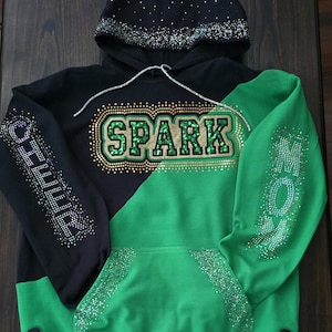 May include: A black and green hoodie featuring the word SPARK in green glitter on the chest, with the word CHEER on the right sleeve and MOM on the left sleeve in rhinestones. The hood and sleeves are accented with rhinestones and black bows.