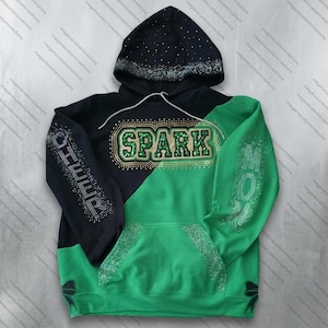 Könnte beinhalten: Ein schwarz-grüner Kapuzenpullover mit dem Wort SPARK in grünen Glitzerbuchstaben auf der Brust, umgeben von goldenen Strasssteinen. Das Wort CHEER steht auf dem rechten Ärmel und MOM auf dem linken Ärmel in Strass. Die Kapuze ist mit Strass besetzt.