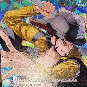 Peut inclure: Une carte à collectionner One Piece holographique représentant le personnage Dracule Mihawk avec un chapeau à larges bords, brandissant une épée au-dessus de sa tête. La carte affiche du texte incluant 5000 de puissance et des instructions.