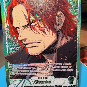May include: A holographic One Piece trading card featuring the character Shanks with vibrant red hair and a stern expression. The card displays a power level of 5000, the text Leader Shanks, The Four Emperors/Red-Haired Pirates, and game instructions.