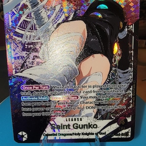 May include: A holographic trading card featuring the character Saint Gunko with blue hair, goggles, and a black outfit. The card displays text including Once Per Turn, Activate:Main, and Celestial Dragons/Holy Knights of God with a power of 5000.