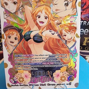 Puede incluir: Una tarjeta coleccionable con una ilustración central de un personaje de pelo largo naranja con top de bikini, rodeado de retratos fragmentados. La tarjeta muestra el texto "LEADER NAMI" y "5000" con las reglas del juego impresas debajo.
