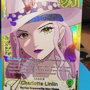 May include: A holographic One Piece trading card featuring Charlotte Linlin as a leader. The card displays a character with long pink hair, wearing a white hat with a purple polka-dot band. Text includes 'When Attacking', 'Charlotte Linlin', and 'The Four Emperors/Big Mom Pirates' with a power level of 5000 and 5 life points.