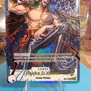 May include: A holographic One Piece trading card featuring the character Rocks D. Xebec. The card displays the text When Attacking You may trash 1 card from your hand: Reveal 1 card from the top of your deck. If the revealed card's type includes Rocks Pirates, draw 2 cards.