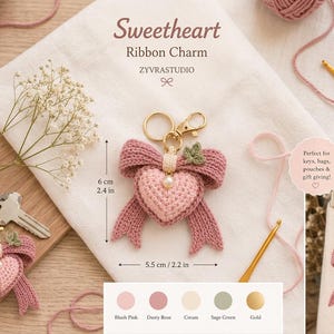 May include: A crochet heart and ribbon charm with a gold-tone lobster clasp. The heart is light pink, the ribbon is dusty rose, and it features a small cream pearl and a sage green leaf accent. It measures 6 cm by 5.5 cm, or 2.4 inches by 2.2 inches.
