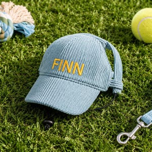 May include: A light blue corduroy baseball cap featuring the name FINN embroidered in yellow thread on the front panel. The cap includes a black adjustable chin strap and side openings for ears, designed with a textured fabric finish.
