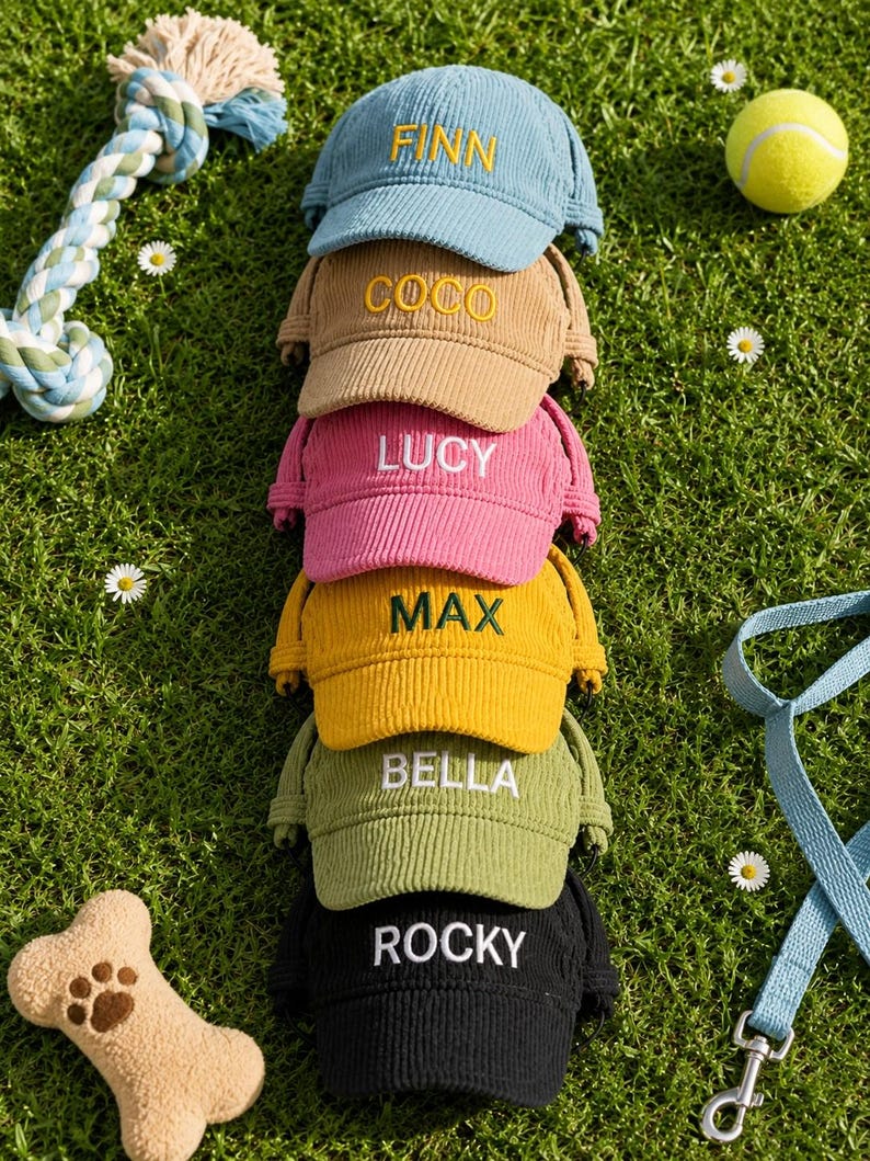 May include: A stack of six corduroy baseball caps for dogs in blue, tan, pink, yellow, olive green, and black. Each cap features an embroidered name on the front: FINN, COCO, LUCY, MAX, BELLA, and ROCKY. The caps have side ear openings.