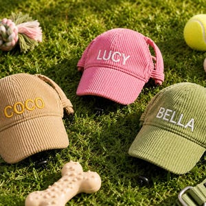 May include: Three corduroy baseball caps for pets in pink, tan, and olive green. The pink cap features the embroidered name LUCY, the tan cap features COCO in yellow, and the olive green cap features BELLA in white. Each cap has side ear holes.