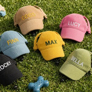May include: A collection of six corduroy pet caps in various colors, including tan, pink, blue, yellow, black, and green. Each cap features an embroidered name on the front: COCO, LUCY, FINN, MAX, ROCKY, and BELLA, with adjustable straps.