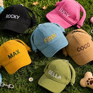 May include: Six corduroy baseball caps for pets in black, pink, blue, yellow, tan, and olive green. Each cap features an adjustable strap and is embroidered with a name: ROCKY, LUCY, FINN, MAX, COCO, or BELLA in contrasting thread colours.