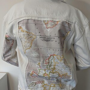 May include: Cream-coloured denim jacket featuring a vintage map design on the back. The map shows continents and oceans in muted tones of beige, brown, and yellow. The jacket has a classic collar, button closures, and long sleeves.