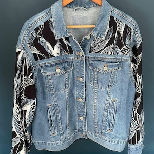 May include: A light blue denim jacket with black sleeves and shoulder panels decorated with a white and black leaf pattern. The jacket has a classic design with button closures, pockets, and a collar, hanging on a wooden hanger.