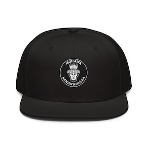 Op de afbeelding: Een zwarte snapback-pet met een cirkelvormige witte afbeelding op het voorpaneel. De afbeelding toont een gestileerd gezicht met een verentooi, omringd door de tekst MOHAWK en KANIEN'KEHÁ:KA in een cirkelvormig randontwerp.