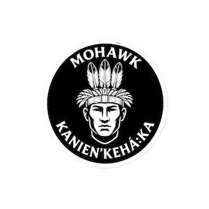 Op de afbeelding: Een ronde zwart-witte sticker met een gestileerde illustratie van een persoon die een traditionele verentooi draagt. De tekst MOHAWK staat in een boog bovenaan en KANIEN'KEHÁ:KA staat in een boog onderaan het cirkelvormige ontwerp.
