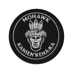 May include: A circular embroidered patch with a black background and white stitching. The design features a central portrait of a person wearing a feathered headdress, with the text MOHAWK arched above and KANIEN'KEHÁ:KA arched below the figure.