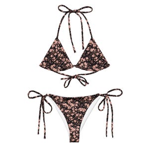 May include: A two-piece bikini set featuring a black fabric base with a small pink floral pattern. The top has triangular cups with thin tie-up straps at the neck and back. The bottom features side tie-up closures and a white interior lining.