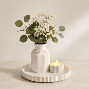 Puede incluir: Un jarrón de cerámica blanca acanalada que contiene un ramo de hojas de eucalipto verdes y flores de paniculata blancas, acompañado de un pequeño portavelas acanalado a juego con una vela LED encendida, sobre una bandeja redonda acanalada.